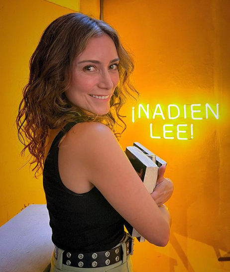 Ana María, de espalda pero mirando a la cámara, sostiene libros con sus brazos. Al fondo, en una pared amarilla, escrita con luz de neón, se ve la frase “¡Nadien lee!”