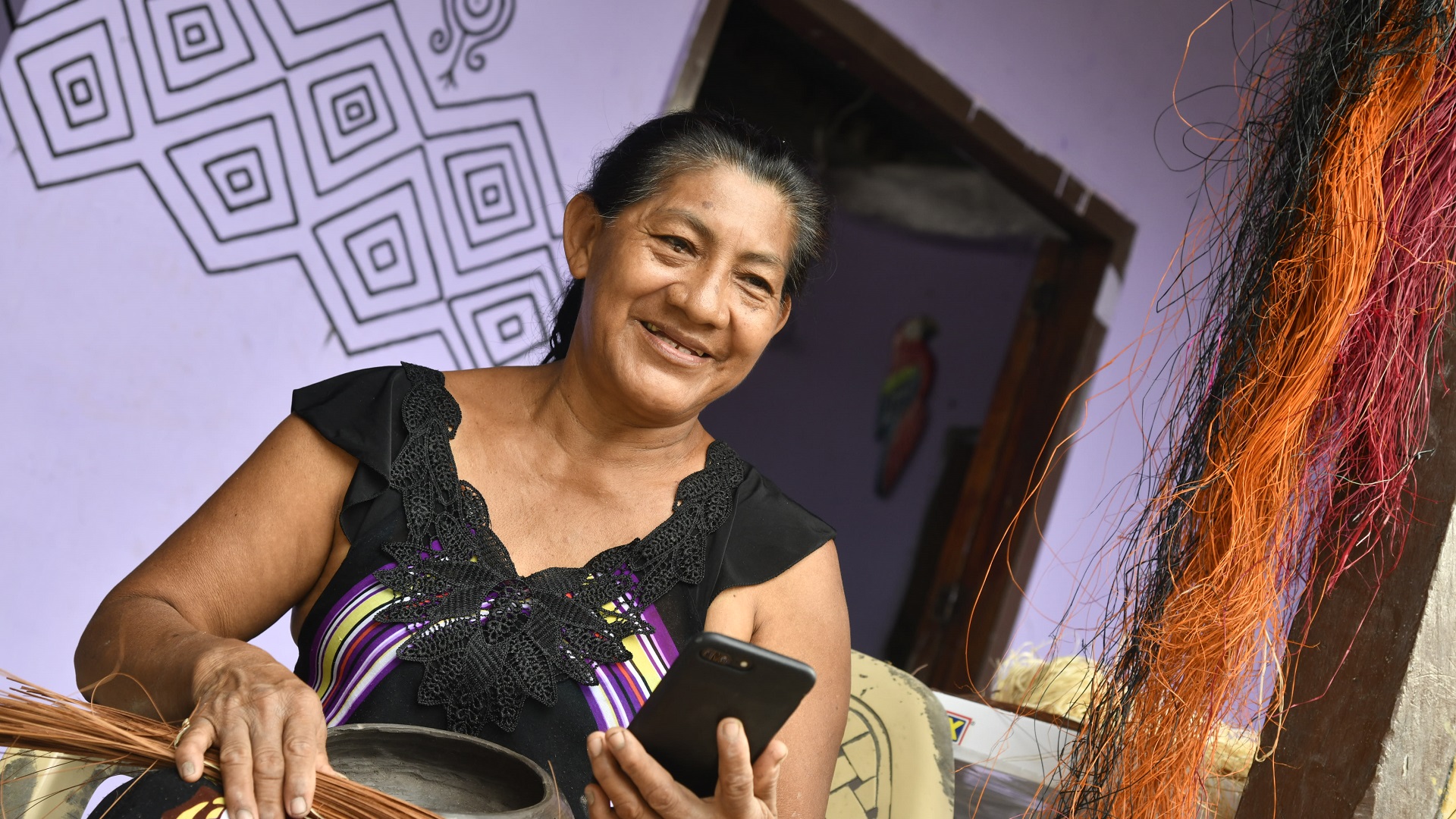 Una mujer haciendo una artesania mientras sostiene un dispositivo movil en su mano izquierda